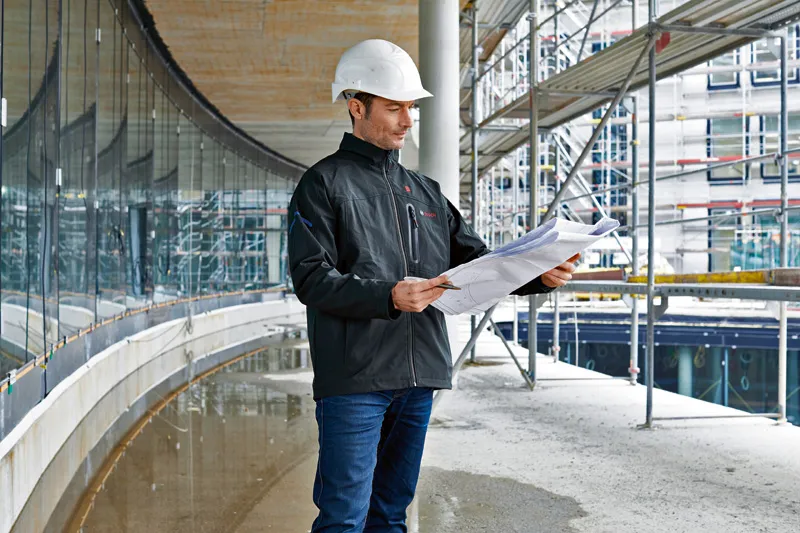 Eine Person mit Sicherheitsausrüstung und beheizter Jacke überprüft auf einer Baustelle die Baupläne.