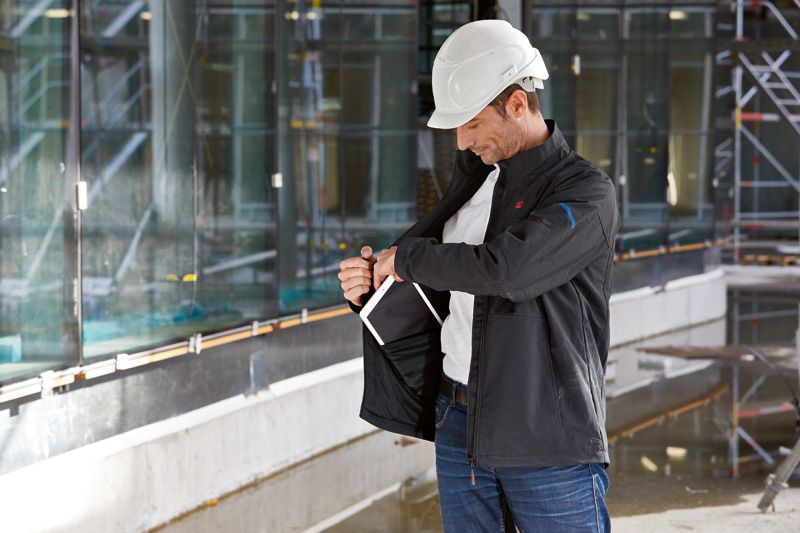 Eine Person mit Sicherheitsausrüstung passt auf einer Baustelle eine beheizte Jacke an.
