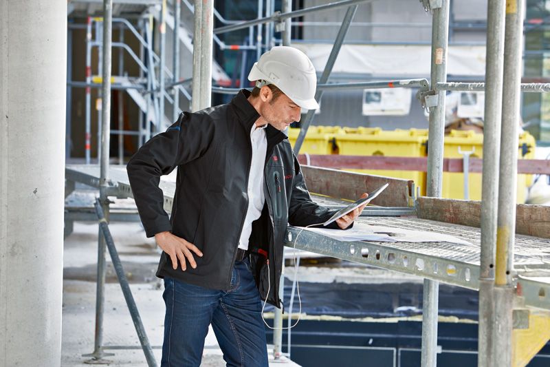 Eine Person mit Sicherheitsausrüstung und beheizter Jacke inspiziert auf einer Baustelle ein Tablet.