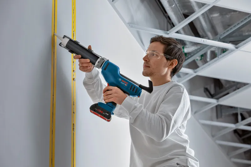 Eine Person mit Sicherheitsausrüstung trägt mit einer kabellosen Kartuschenpistole Dichtungsmittel auf eine Wand auf.