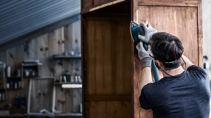 Eine Person mit Schutzausrüstung schleift einen Holzschrank mit einem Elektrowerkzeug.