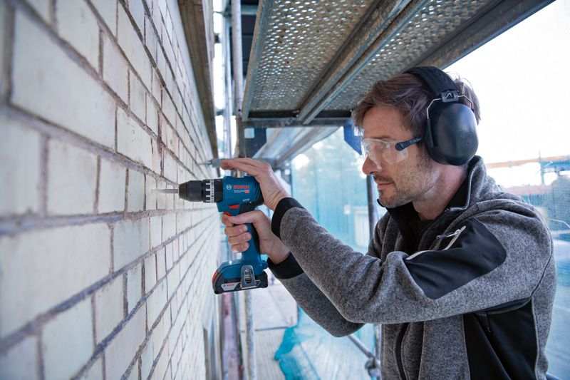 Person mit Schutzausrüstung bohrt mit einem Akku-Elektrowerkzeug in eine Ziegelwand.