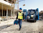 Ein Arbeiter in Sicherheitsausrüstung trägt einen Werkzeugkoffer neben einem mit Elektrowerkzeugen vollgestopften Lieferwagen.