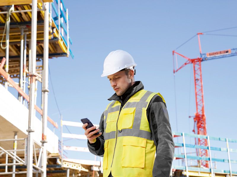 Eine Person mit Sicherheitsausrüstung überprüft auf einer Baustelle ein Smartphone.