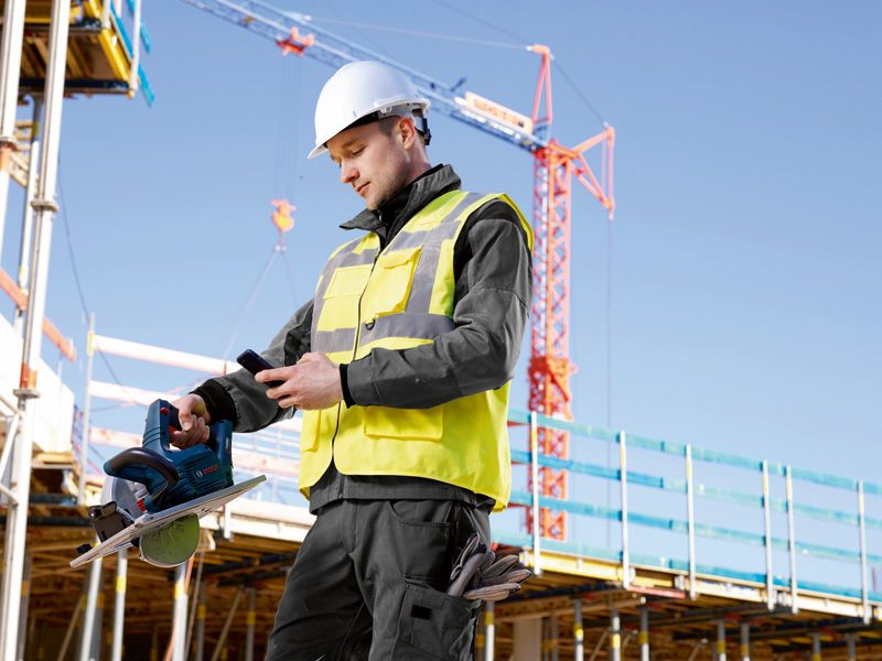 Eine Person mit Sicherheitsausrüstung überprüft auf einer Baustelle ein Telefon, während sie eine Kreissäge hält.