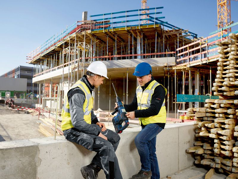 Zwei Personen in Sicherheitsausrüstung besprechen auf einer Baustelle einen Bohrhammer.