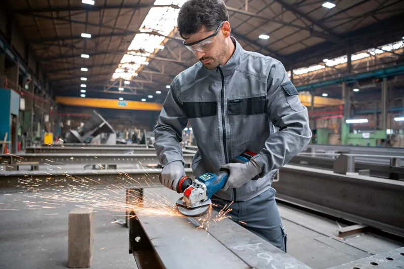 Eine Person, die Schutzausrüstung trägt, schleift einen Metallträger mit einem Akku-Winkelschleifer.