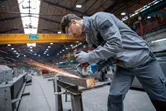 Eine Person in Schutzausrüstung schleift einen Metallträger in einer Industriewerkstatt.
