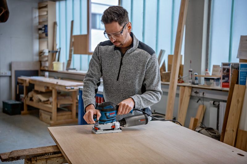 Eine Person mit Schutzausrüstung schleift ein Holzbrett mit einem Schwingschleifer.