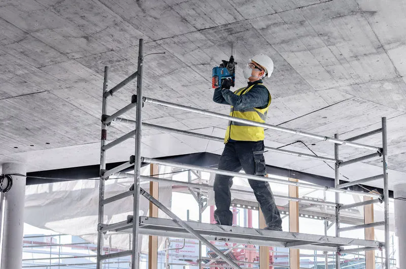 Eine Person mit Sicherheitsausrüstung bohrt auf einem Gerüst stehend in eine Betondecke.