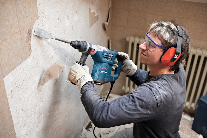 Eine Person mit Schutzausrüstung entfernt mit einem Bohrhammer Fliesen aus einer Wand.