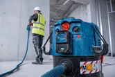 Eine Person, die Schutzausrüstung trägt, bedient ein Elektrowerkzeug, das an eine Staubabsaugung angeschlossen ist.