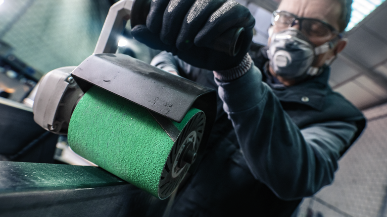 Eine Person mit Schutzausrüstung poliert eine Metalloberfläche mit einem elektrischen Schleifgerät.