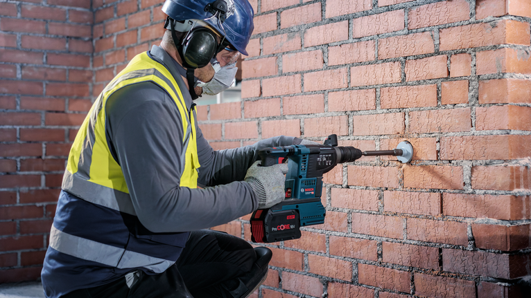 Person mit Sicherheitsausrüstung bohrt mit einem Elektrowerkzeug in eine Ziegelwand.