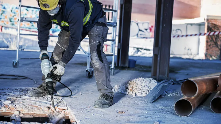 Eine Person mit Sicherheitsausrüstung bohrt auf einer Baustelle durch Betonschutt.