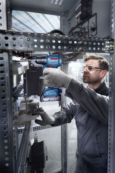 Eine Person mit Sicherheitsausrüstung verwendet eine Akku-Bohrmaschine in einem Schaltschrank.