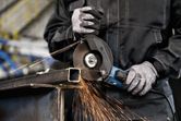 Eine Person mit Schutzausrüstung schleift mit einem Winkelschleifer einen Metallträger, wodurch Funken entstehen.