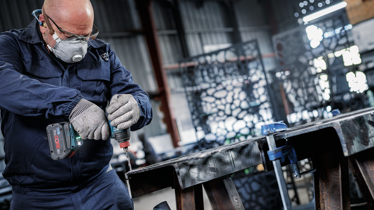 Eine Person mit Sicherheitsausrüstung bohrt in einer Industriewerkstatt in einen Metallträger.