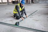 Eine Person, die Schutzausrüstung trägt, richtet mithilfe eines Laser-Nivelliergeräts Metallschienen auf einer Baustellenfläche aus.