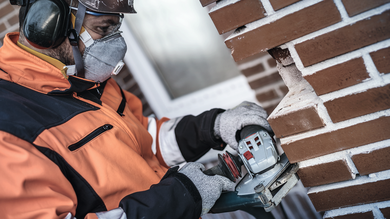Ein Arbeiter mit Schutzausrüstung schneidet mit einem Elektrowerkzeug Rillen in eine Ziegelwand.