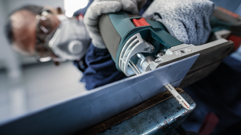 Eine Person mit Sicherheitsausrüstung schneidet mit einer Stichsäge ein Metallblech.