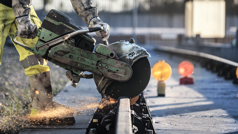 Eine Person, die Schutzausrüstung trägt, schneidet mit einem Elektrotrennschleifer eine Metallschiene durch, wobei Funken entstehen.