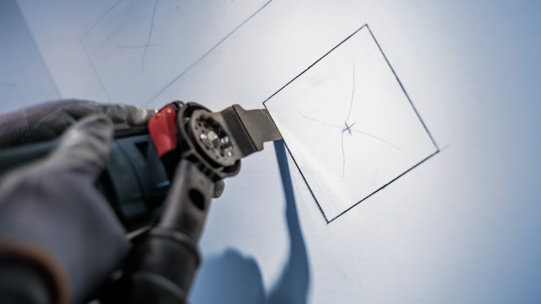 Eine Person mit Sicherheitsausrüstung schneidet mit einem Elektrowerkzeug ein Quadrat in eine Trockenbauwand.