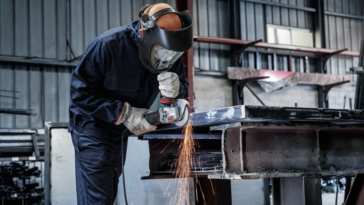 Eine Person mit Sicherheitsausrüstung schleift in einer industriellen Umgebung Metall und erzeugt dabei Funken.