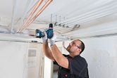 Eine Person mit Sicherheitsausrüstung bohrt in eine Decke, um elektrische Leitungen zu installieren.