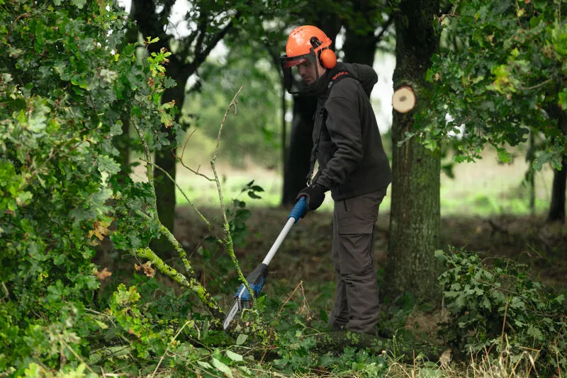 Eine Person mit Schutzausrüstung schneidet Äste mit einer kabellosen Teleskop-Gartenschere.