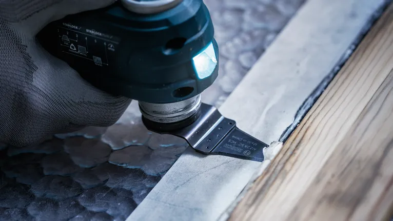 Eine Person, die Schutzausrüstung trägt, benutzt ein Multifunktionswerkzeug, um Material neben Holzleisten zu schneiden.