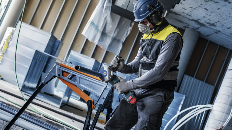 Eine Person mit Sicherheitsausrüstung bohrt auf einer Baustelle in ein Metallrohr.