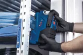 Eine Person in Schutzausrüstung installiert ein blaues Elektrowerkzeug in der Nähe von Metallregalen.