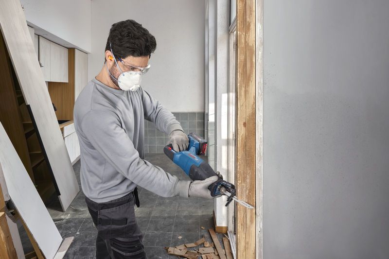 Eine Person, die Schutzausrüstung trägt, benutzt eine akkubetriebene Säbelsäge, um einen Holzrahmen zu zersägen.