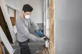 Eine Person, die Schutzausrüstung trägt, benutzt eine akkubetriebene Säbelsäge, um einen Holzrahmen zu zersägen.