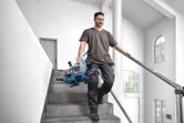 Eine Person, die Schutzausrüstung trägt, trägt eine Gehrungssäge, während sie eine Treppe hinuntergeht.