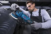 Eine Person in Schutzausrüstung poliert ein schwarzes Auto mit einer elektrischen Poliermaschine.