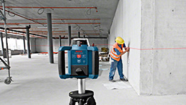 Bosch Professional Lasergerät im Vordergrund, Arbeiter mit gelber Helm an Wand.