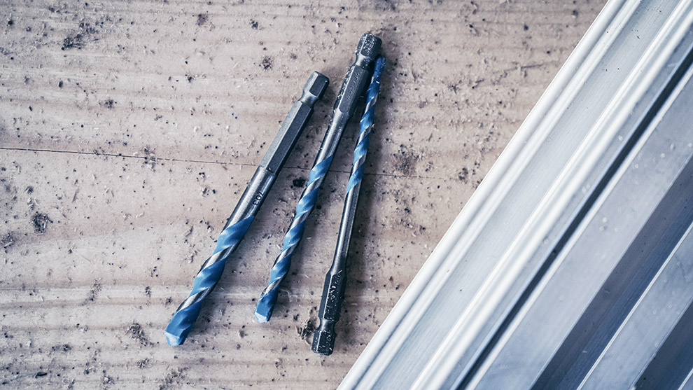 Drei blaue Bosch Professional Bohrer liegen auf einem Holzuntergrund neben Metallprofilen.