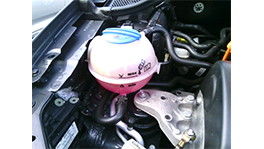 Ein pinker Kühlmittelbehälter im Motorraum eines Autos, umgeben von Schläuchen und Metallteilen.