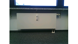 Ein weißer Heizkörper unter einem Fenster, auf einem dunklen Teppichboden.