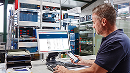 Ein Mann arbeitet am Computer in einem Lager mit Werkzeugregalen im Hintergrund.