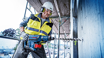 Ein Arbeiter mit Gehörschutz bohrt mit einem blauen Bosch Professional Werkzeug in eine Wand.