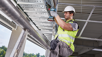 Ein Handwerker in gelber Warnweste befestigt Metallprofile mit einem Akkuschrauber.