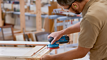 Ein Handwerker schleift eine Holzplatte mit einem blauen Bosch Professional Schleifer.
