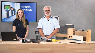 Zwei Personen stehen an einem Tisch mit Werkzeugen und Holzmaterialien, Bildschirm im Hintergrund.