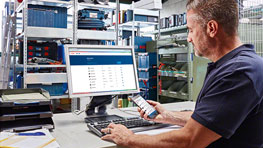 Mann am Computer in Werkstatt, hält Smartphone, umgeben von Werkzeugregalen.