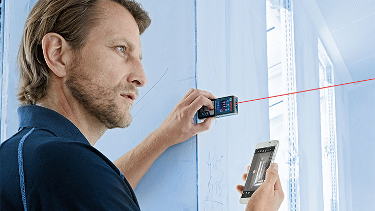 Ein Mann nutzt einen Bosch Professional Laser-Entfernungsmesser an einer blauen Wand.