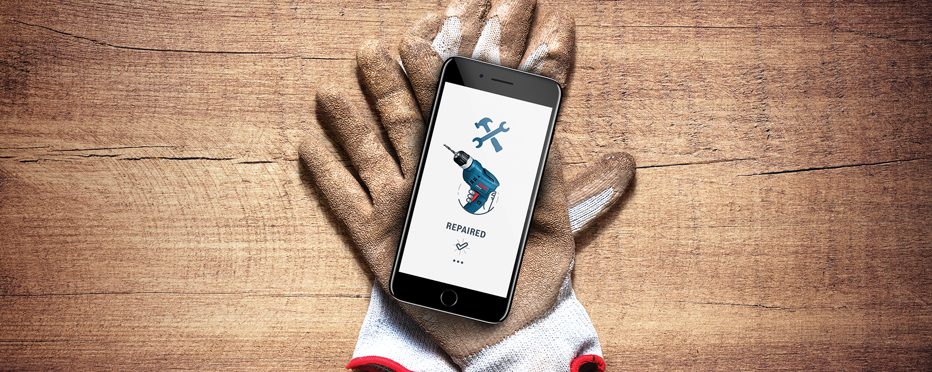 Ein Smartphone auf Arbeitshandschuhen zeigt ein Werkzeug-Icon auf Holzuntergrund.