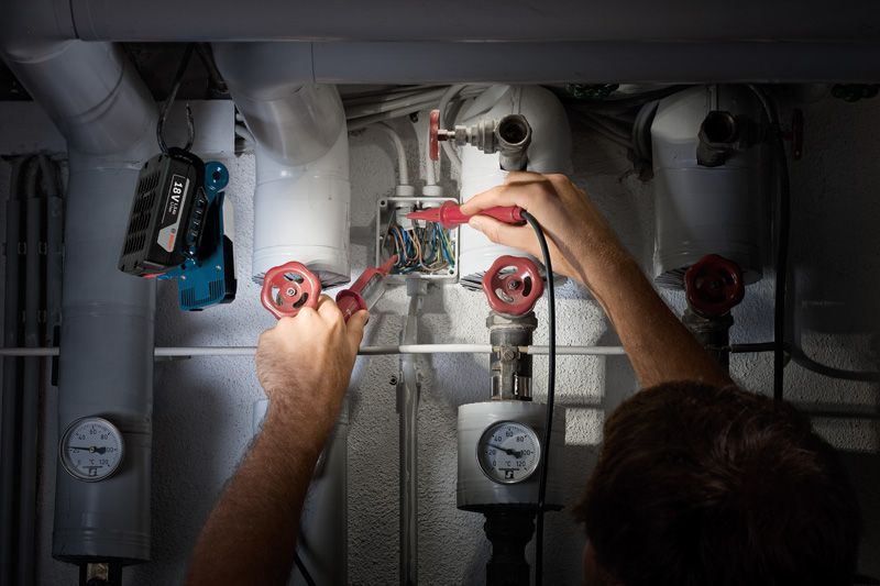 Person tjekker elektriske ledninger nær rør, ledningsfri arbejdslampe monteret ovenfor.